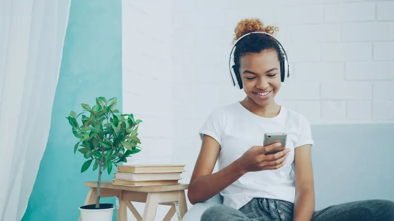 Smiling young person with headphones looking at a phone in a bright living room