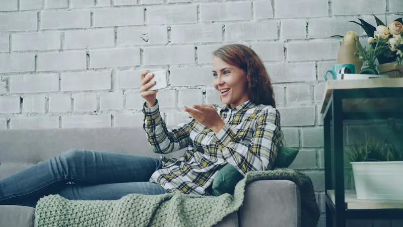 Smiling woman using her phone, reclining on a gray sofa with a blanket